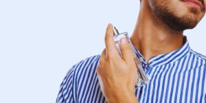 Homem de camisa azul e branca espirrando perfume no próprio pescoço