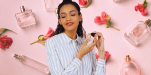 Mulher sorridente passando perfume; ao fundo, montagem com frascos de perfumes e flores